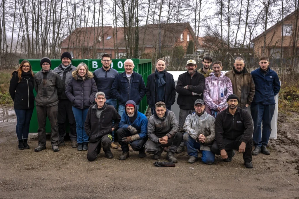 Photo de l'équipe de Beniva - 17 collaborateurs, ouvriers, soudeurs, caristes, peintre, laveurs, fonctions supports et direction. L'équipe de Beniva, est photographiée à l'extérieur sur leur site, devant une benne ampliroll - produit phare que conçoit l'entreprise Beniva en France, dans leur atelier à Lure